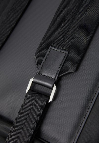 Black fabric backpack strap featuring a textured surface, leather accents, and a metallic rectangular buckle for adjustment.