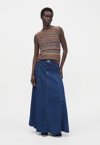 Femme portant un haut rayé multicolore sans manches et une longue jupe en jean bleue, debout devant un fond blanc uni.