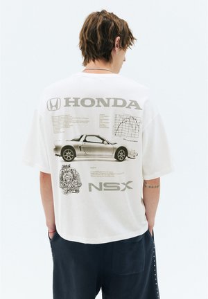 Personne aux cheveux courts portant une chemise blanche avec une illustration de la voiture Honda NSX et des détails techniques au dos, tournée dos à la caméra.