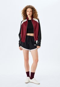 Burgundowa i czarna wiatrówka z białymi akcentami, krótki krój; zestawiona z czarnymi szortami z białymi lamówkami i logo, noszona z białymi butami.