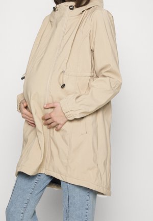 Personne enceinte portant une veste de maternité beige, les mains reposant sur le ventre sur un fond uni.
