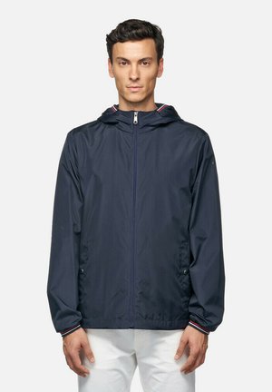 Chaqueta ligera azul marino con capucha, con cremallera frontal y dos bolsillos; detalles a rayas rojas y blancas en el cuello.