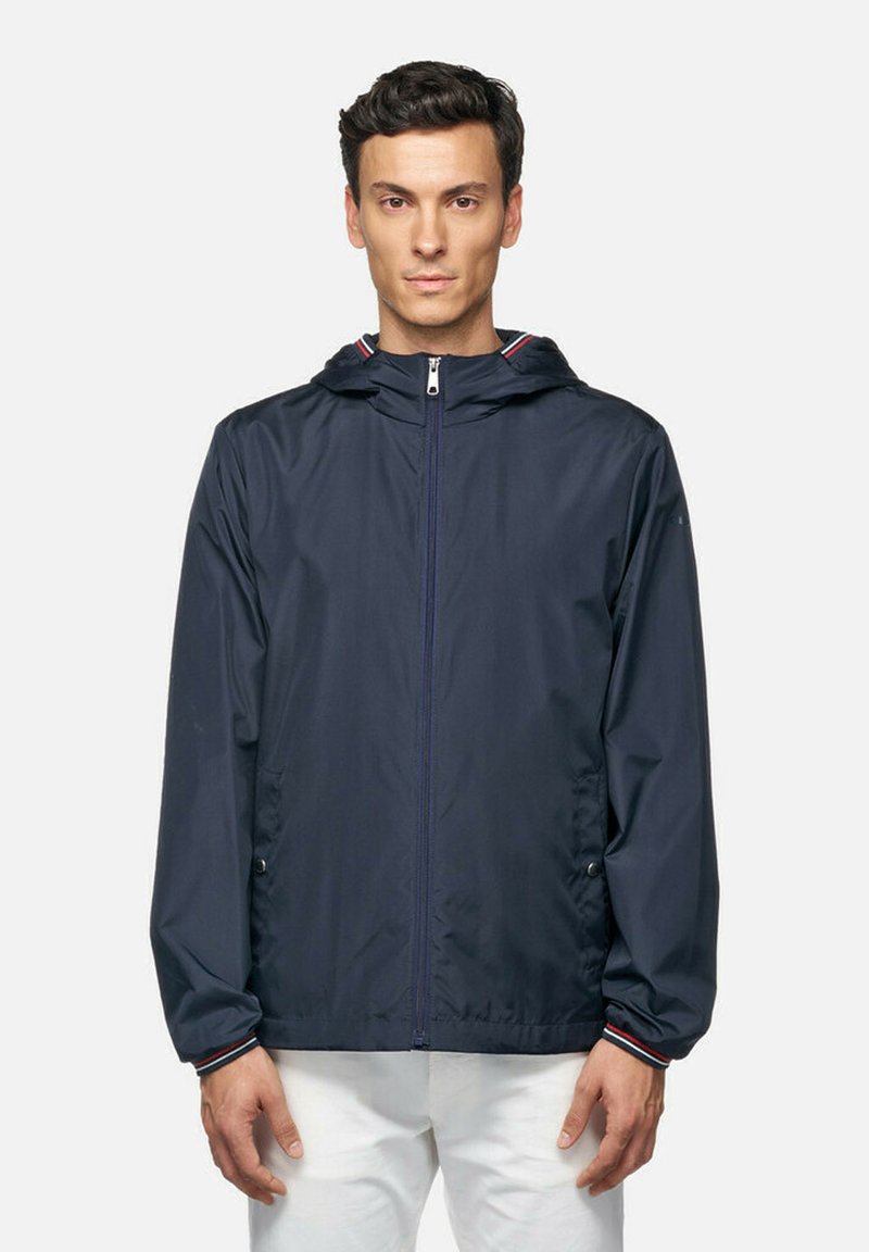Marineblauwe lichtgewicht jacket met capuchon, voorzien van een voorsluiting met rits en twee zakken; rode en witte gestreepte accenten op de kraag.