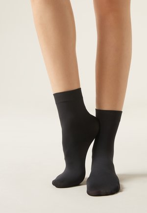 Svarta ankelsockor av ett slätt, elastiskt material med ribbad mudd och en sömlös design, placerade på bara ben mot en ljus bakgrund.