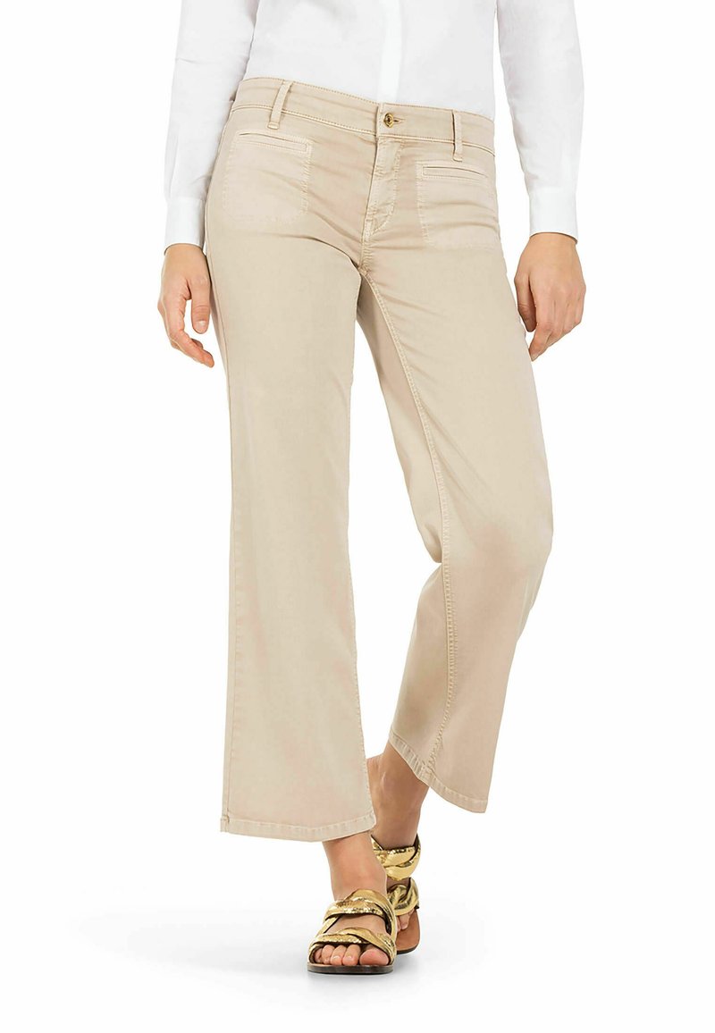 Beige weite Hosen mit vorderen Taschen, einem Knopfverschluss und einer glatten Textur, kombiniert mit Sandalen mit goldenen Riemen.