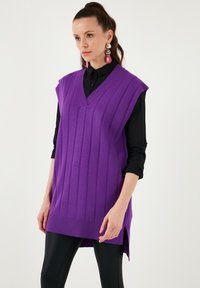 Purple sleeveless knit sweater with a V-neck, featuring vertical ribbed texture and side slits. Worn over a black button-up shirt.
