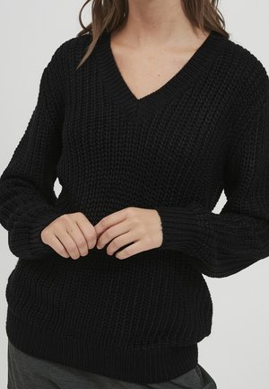 Femme portant un pull en maille épaisse noir avec un col en V et des manches longues, les mains jointes près de la taille sur un fond uni.