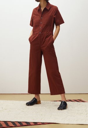 Rdzawy, krótko rękawowy kombinezon z dwiema kieszeniami na piersi i szerokimi nogawkami. Połączony z czarnymi plecionymi klapkami na jasnym, teksturowanym dywanie.