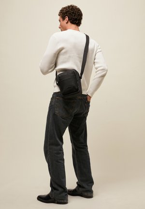 Mees, kes kannab valget kampsunit ja tumedaid teksaseid, kannab väikest musta ristihoidjat, seisab ühevärvilise beežitausta ees, küljele näoga.