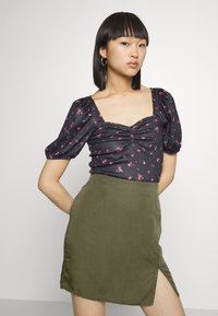 Top preto floral com mangas bufantes e decote quadrado, emparelhado com uma saia verde com uma fenda lateral e textura de tecido suave.