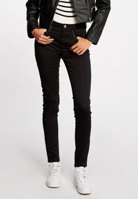 Vrouw in zwarte slim-fit broek, witte sneakers, gestreepte top en een kort zwart leren jack, staande met haar handen in haar zakken.