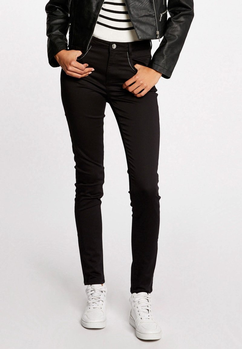 Vrouw in zwarte slim-fit broek, witte sneakers, gestreepte top en een kort zwart leren jack, staande met haar handen in haar zakken.