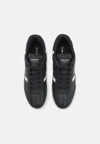 Black sneakers with a textured upper featuring geometric patterns, white side stripes, and black laces. Lacoste branding on the tongue.
