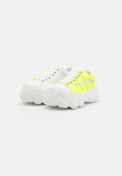 Λευκά και νέον κίτρινα αθλητικά παπούτσια με υφή στην επάνω πλευρά, στρογγυλή πλατφόρμα και ζιγκ-ζαγκ σχέδιο. Διαθέτουν μεταλλικές θηλιές και κορδόνια.