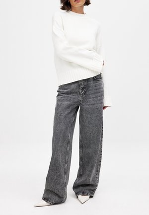 Kobieta ubrana w biały, oversize'owy sweter, luźne ciemnoszare jeansy z efektem sprania i białe, szpiczaste buty, stojąca na jednolitym tle.