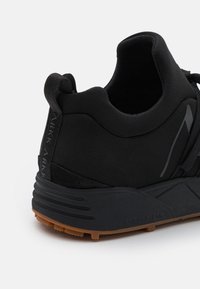 Czarny but sportowy z teksturowaną górą z tkaniny, amortyzowaną podeszwą i kontrastową brązową gumową podeszwą zewnętrzną. Posiada pętelkę u pięty.