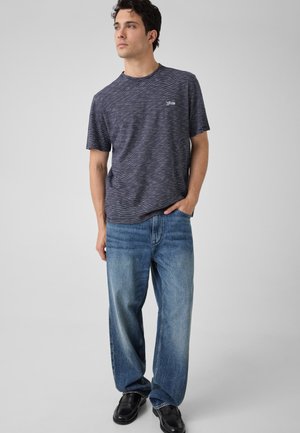 Jeune homme debout, main gauche dans la poche, portant une chemise à manches courtes rayée foncée, un jean bleu ample et des chaussures noires, regardant vers la droite.