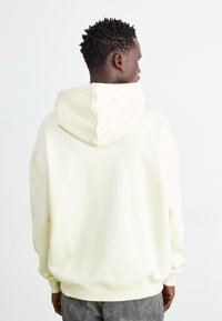 Cremefarvet hoodie med afslappet pasform, der har en stor hætte og ribbede manchetter. Stoffet har en glat overflade og minimal detaljering.