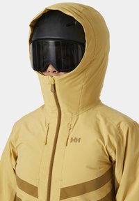 Gelbe isolierte Jacke mit hohem Kragen, Reißverschluss und braunen horizontalen Streifen; getragen mit einem schwarzen Skihelm und getönten Skibrillen.