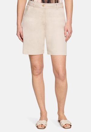 Vrouw draagt beige knielange shorts met voorzakken en knoopsluiting, gecombineerd met beige geweven slidesandalen.