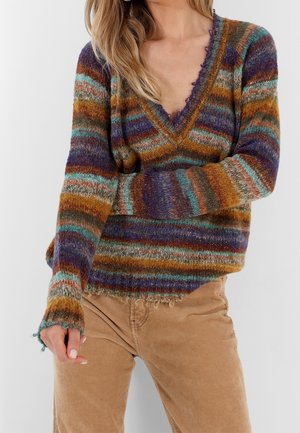 Femme portant un pull duveteux rayé multicolore à col en V et un pantalon en velours côtelé beige sur un fond uni.