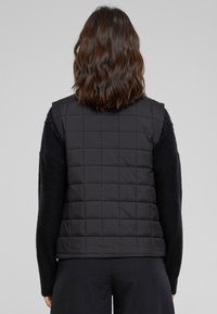 Gilet matelassé noir avec un col montant et sans manches, fabriqué en tissu lisse, présentant une couture carrée et une coupe ajustée.