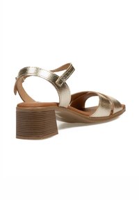 Sandalias metálicas doradas con tacón cuadrado, que presentan un diseño entrecruzado, una correa de tobillo ajustable y una plantilla texturizada.