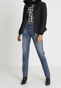 Svart puffjacka, mönstrad svart och vit topp, blå jeans med fransad fåll, matchat med ankelboots, står framför en neutral bakgrund.