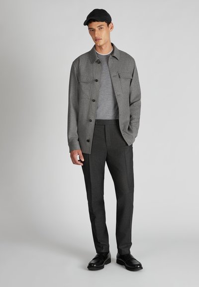 Graue Hemdjacke über einem grauen Pullover, kombiniert mit dunklen Hosen und schwarzen Schuhen. Zu den Merkmalen gehören zwei Vordertaschen und eine Mütze.
