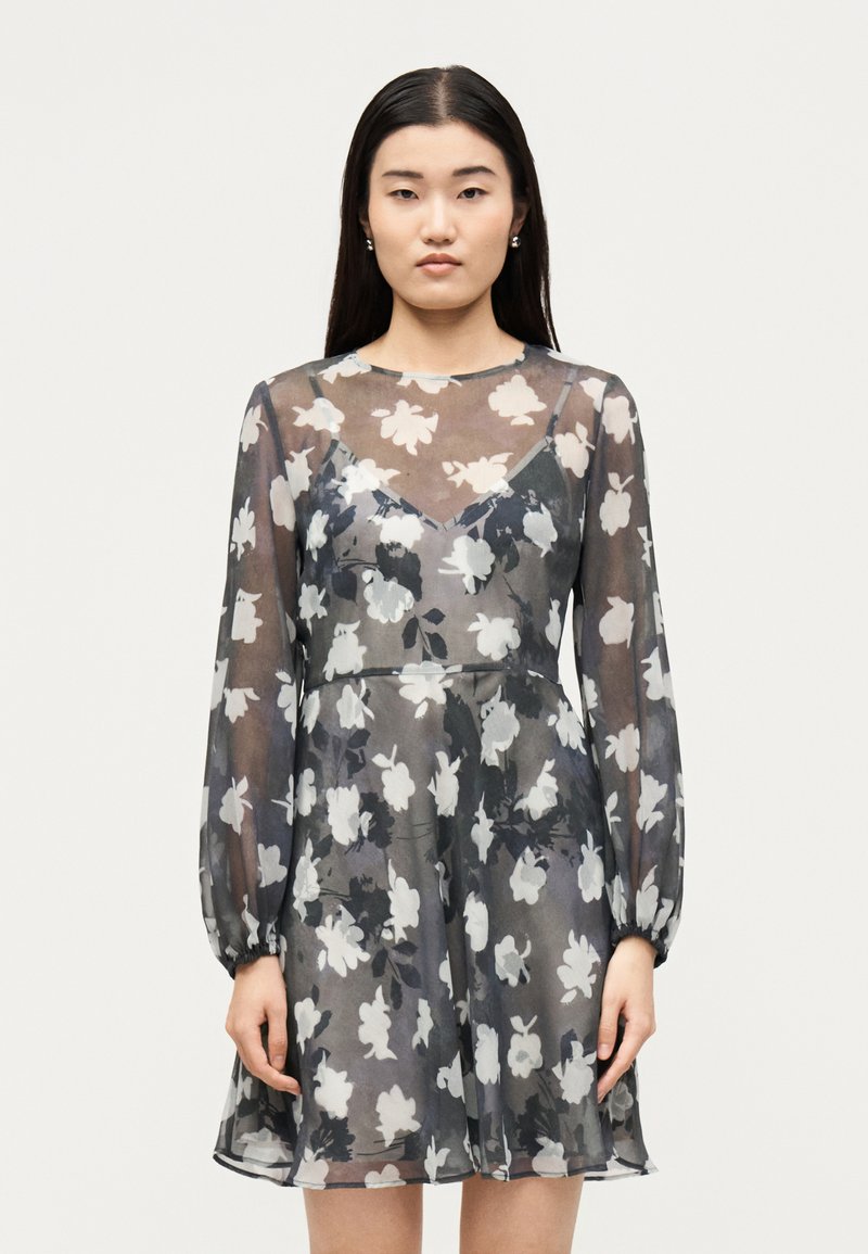 Γυναίκα φοράει μαύρο διαφανές μακρυμάνικο φόρεμα με λευκά λουλουδάτα σχέδια και μαύρο εσώρουχο από κάτω, στέκεται μπροστά σε μονόχρωμο φόντο.