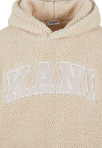 Sweat à capuche en polaire sherpa couleur crème avec de grandes lettres blanches en contour "KANI" sur la poitrine.