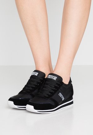 Gambe con indosso sneakers nere Versace Jeans Couture con suole bianche, design con lacci e patch con logo sulla linguetta e sul lato.