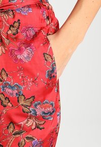 Röd satin tyg med blommönster i blått, rosa och guld. Har en sidficka och en slät, glänsande textur.