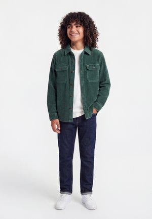 Jong persoon glimlacht, draagt een groen corduroy overhemd over een wit t-shirt, donkere jeans en witte sneakers, staat met één hand in de zak.