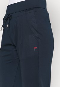 Marinblå sportbyxor med dragsko i midjan, sidofickor och en liten logotyp i rött och grått på vänster lår. Slät textur.