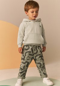Hellgraues Hoodie mit einer Vorderentasche und einem kleinen Logo, kombiniert mit grünen gemusterten Cargo-Hosen und weißen Sneakern. Glatte Textur.