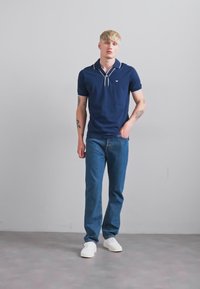 Tânăr cu păr blond, purtând un tricou polo bleumarin, blugi și adidași albi, stând în fața unui zid simplu de culoare gri deschis.