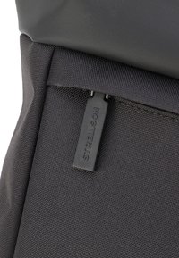 Détail d'un sac à dos en tissu noir mettant en avant un matériau texturé et une tirette de zip avec le nom de la marque "Strellson" embossé dessus.