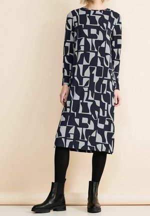 Nainen, joka pukeutunut musta-harmaaseen abstraktikuviolliseen pitkähihaiseen midimekkoon, mustiin leggingsseihin ja mustiin nilkkasaappaisiin, seisomassa beigenväristä seinää vasten.