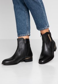 Bottes cheville en cuir noir avec fermeture éclair, talon bas carré et détail accent doré. Texture lisse et design à bout pointu.