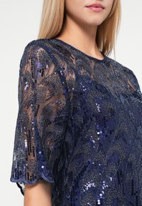Blouse bleu marine en tissu transparent ornée de sequins. Motif floral avec des détails texturés et des manches à bords festonnés. Encolure ronde.