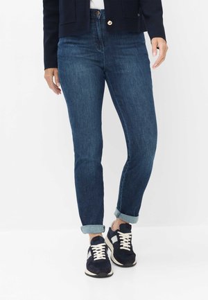 Personne portant un jean bleu foncé retroussé, des baskets bleu marine avec des lacets blancs et une veste bleu marine, debout devant un fond blanc.