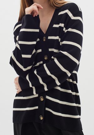 Kvinde iført sort cardigan med hvide vandrette striber og store knapper, armene krydset, hånd nær ansigtet, ensfarvet lys baggrund.