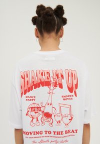 Hvid oversized t-shirt med fed rød grafisk tekst og tegneserieelementer, der viser danse-temaer og karakterer på ryggen.
