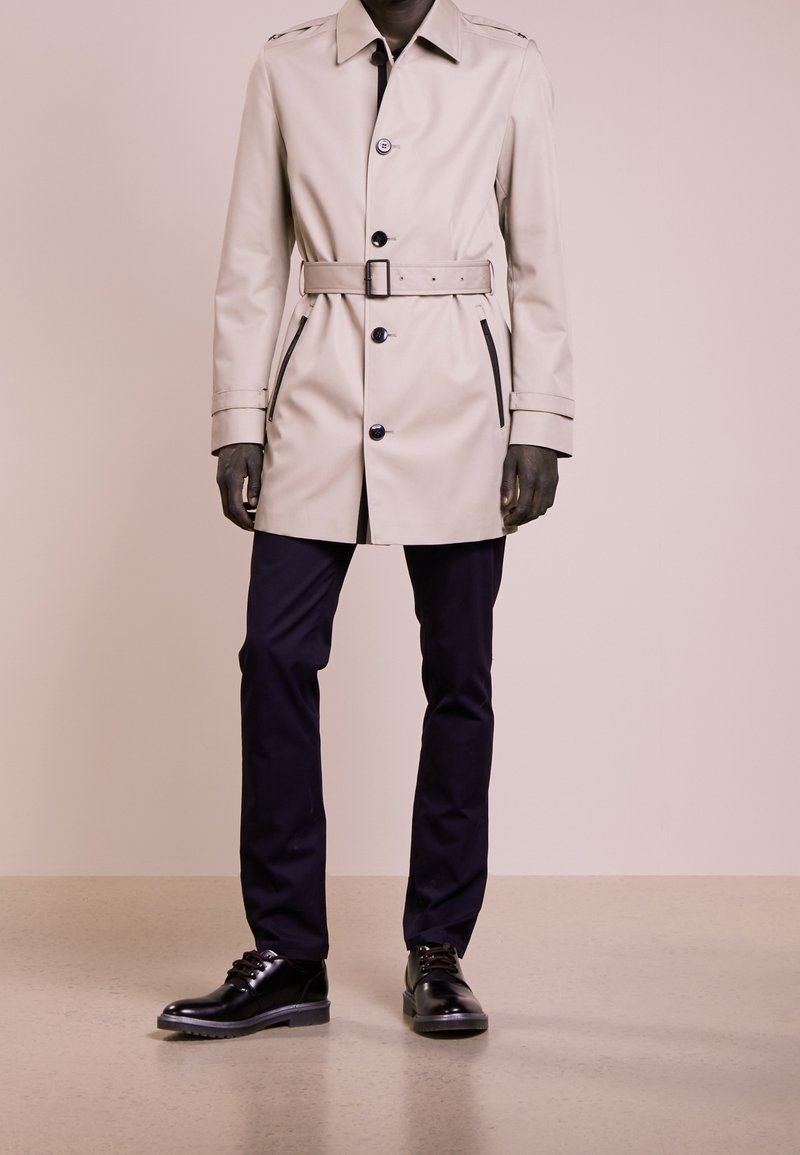 Trench coat beige con vita regolabile con cintura, chiusura a bottoni e tasche laterali, abbinato a pantaloni blu navy e scarpe nere stringate su un pavimento neutro.