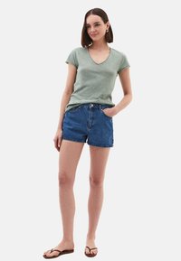 T-shirt verde a scollo a V, pantaloni corti di jeans e infradito. La maglietta è leggera con una vestibilità morbida. I pantaloni corti sono blu con un taglio classico e tasche.
