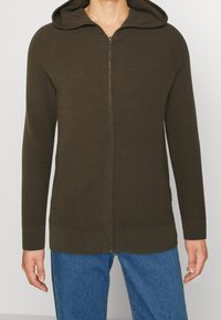 Mörkgrön zip-up hoodie i en strukturerad stickad material, långa ärmar och en justerbar huva, kombinerad med blå denimjeans.