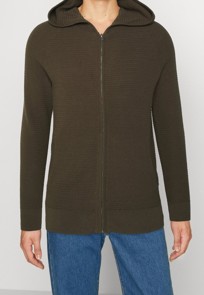 Mörkgrön zip-up hoodie i en strukturerad stickad material, långa ärmar och en justerbar huva, kombinerad med blå denimjeans.