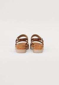 Brown leather sandals with adjustable straps, textured cork-like heel, and a light-colored platform. Back view showcases strap design.