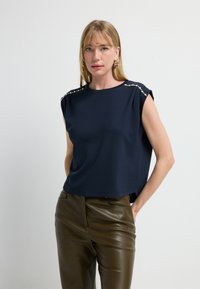 Marineblaues Kurzarmoberteil mit dekorativen silbernen Nieten auf den Schultern, aus weichem Stoff, kombiniert mit eng anliegenden olivgrünen Lederhosen.